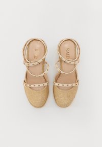 Sandalias de esparto beige con punta de yute trenzado, detalles de tiras ajustables y tachuelas en tono dorado. Interior suave con logo de la marca.