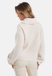 Lichtroze gebreide trui met een brede kraag, oversized mouwen en een ribbelstructuur, getoond vanaf de achterkant met een relaxte pasvorm.