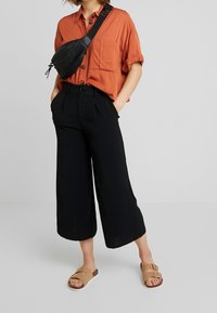 Chemise orange à manches courtes avec boutons et deux poches poitrine, pantalon noir à jambes larges et sandales à glisser beiges avec un détail de boucle.