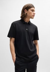 Černé polo tričko vyrobené z hladkého materiálu s klasickým límcem, zapínáním na tři knoflíky a logem na patchi poblíž výstřihu. Krátké rukávy.