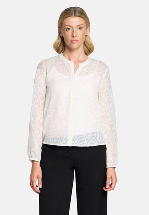 Woman wearing a sheer white long-sleeve blouse with a subtle animal print and black pants, standing against a plain background.