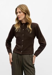 Cardigan marron avec un col haut, des manches longues et des boutons en argent sur le devant. Porté sur un pantalon plissé gris. Texture douce.