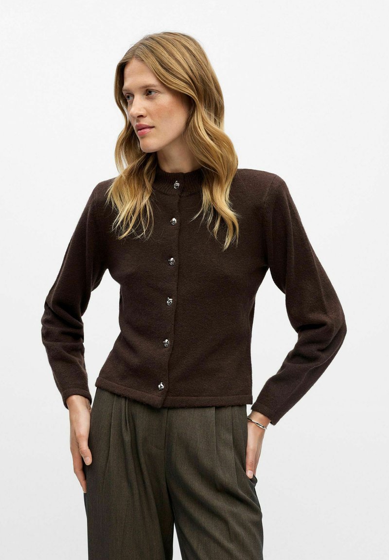 Cardigan marron avec un col haut, des manches longues et des boutons en argent sur le devant. Porté sur un pantalon plissé gris. Texture douce.