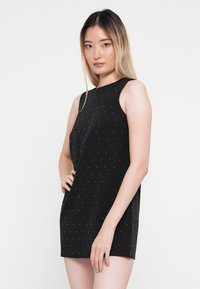 Rochie neagră fără mâneci, cu decolteu rotund, decorată cu un pattern cu puncte de mici cuie argintii, croi drept și textură netedă a materialului.