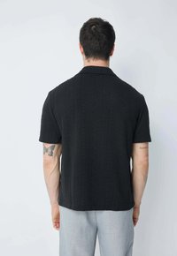 Chemise noire à manches courtes avec des motifs en tricot texturé et un col plat. Le dos présente un ourlet droit et une coupe décontractée.
