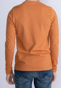 Långärmad orange bomulls t-shirt med rund halsringning, i kombination med blå denimjeans med en bakfickdesign och synliga stygn.