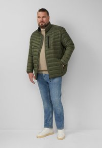 Olivgrüne Steppjacke mit gestepptem Design, hohem Kragen und Reißverschlusstasche. Getragen über einem beigen Pullover und kombiniert mit blauen Jeans und weißen Sneakern.