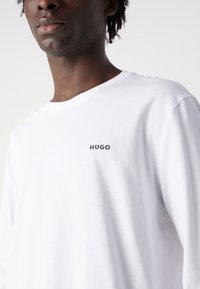 Tricou alb cu mânecă lungă, din bumbac, cu un decolteu rotund și un mic logo negru "HUGO" în zona stângă a pieptului.