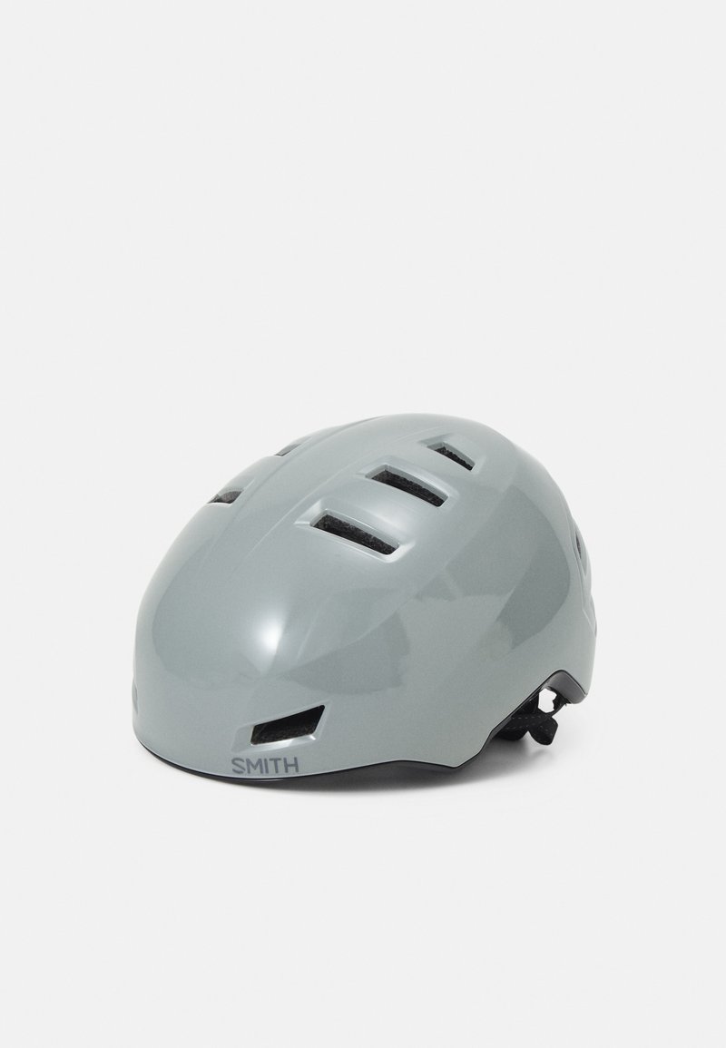Grå cykelhjälm med en slät, glänsande yta, som har flera ventilationsöppningar och ett justerbart spänne. Märkeslogotypen visas på sidan.