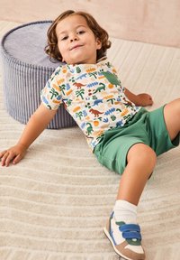 Barn iført en T-shirt med kortærmer og et flerfarvet dinosaurmønster, grønne shorts, hvide sokker og sneakers med blå detaljer, som hviler på et tekstureret tæppe.