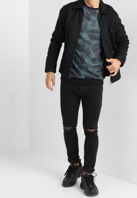 Giacca nera con colletto sopra una camicia verde scuro a motivi con foglie di palma, abbinata a jeans neri strappati e scarpe da ginnastica nere.