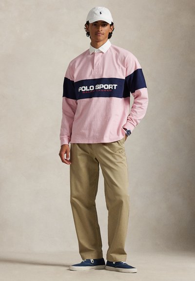 Rózsaszín hosszú ujjú póló tengerészkék csíkokkal és "POLO SPORT" logóval, khaki nadrággal és kék tornacipővel párosítva, fehér sapkával a fején.