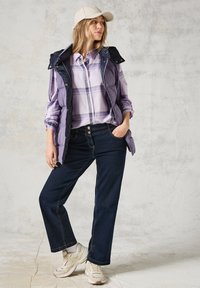 Camisa de cuadros morados debajo de un chaleco acolchado morado y azul marino, combinado con pantalones de mezclilla oscuros y zapatillas claras. Una gorra neutra completa el look.