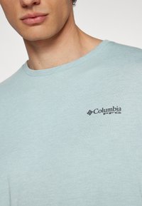 Svetlo modra kratka majica s kratkimi rokavi iz mehkega materiala; vsebuje majhen črn logo Columbia na levem prsnem delu.