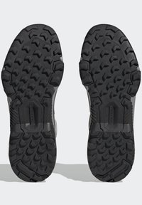 Suole di scarpe in gomma nera con un modello di battistrada profondo. Logo "TRAXION" ben visibile e sezioni testurizzate per una maggiore aderenza e stabilità.