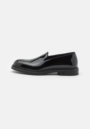 Zapato de charol negro tipo slip-on con punta redondeada, presenta una suela texturizada y un sutil logotipo en el tacón. Acabado de superficie suave.