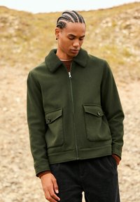 Young man with braided hair wearing a dark green zip-up jacket with large buttoned pockets outdoors on a rocky background.