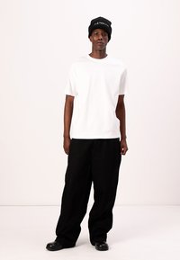 Weißes Baumwoll-T-Shirt, lockere Passform, kombiniert mit schwarzen weiten Hosen und schwarzen Lederschuhen. Schwarze Mütze mit weißem Textakzent.
