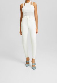 Top corto blanco con ribetes y tirantes cortados, combinado con jeans ajustados blancos de tiro alto y tacones azulados con punta afilada y acentos de hebilla.