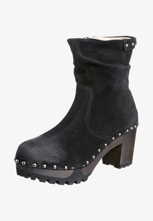 Bottine en daim noir avec bordure cloutée, talon épais et semelle robuste, présentant une tige souple et une sangle décorative avec des clous au sommet.