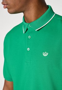 Polo verzi cu un color texturat și accente albe, având trei nasturi și un mic logo alb pe piept.