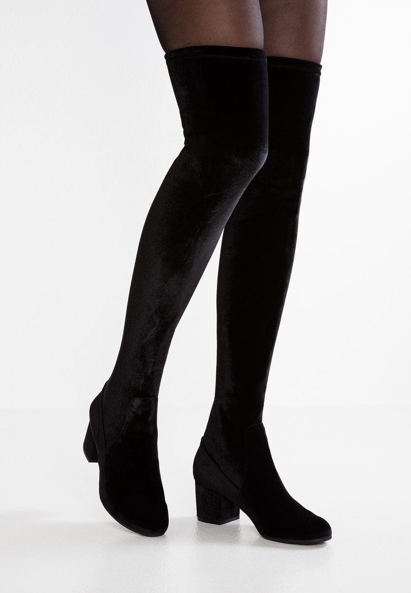 Botas de terciopelo negro por encima de la rodilla con tacones cuadrados, usadas con medias negras transparentes sobre un fondo blanco.
