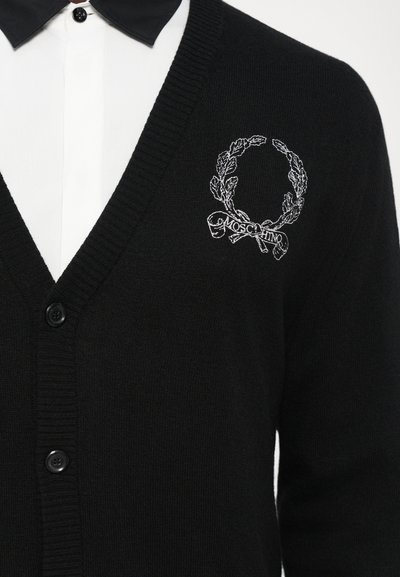 Čierny pletený kardigán s V-výstrihom, ktorý má na ľavej hrudi striebornú vyšívanú vence a logo "Moschino". Päť predných gombíkov.
