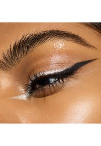 Nahaufnahme eines Auges mit einem markanten schwarzen Eyeliner-Flügel, schimmerndem silbernem Lidschatten und dunklen Wimpern vor einem glatten, gebräunten Augenlid.