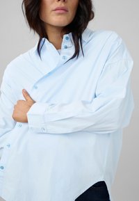 Light blue button-up shirt with a loose fit, contrasting buttons, and a curved hem. The fabric appears smooth and slightly shiny.