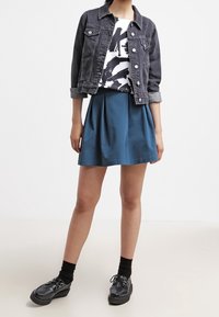 Giacca di denim, camicia bianca con motivo a pennellate nere, gonna plissettata teal, scarpe platform nere e calzini neri alla caviglia.