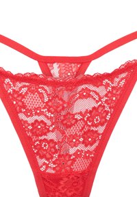 Tanga en dentelle rouge avec motif floral, présentant un devant en dentelle transparente et des bretelles élastiques. Le dos arbore une coupe basse accentuée par de la dentelle.