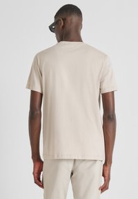 Camiseta de algodón beige con mangas cortas, cuello redondo y un corte relajado, vista desde la parte de atrás. Combinada con pantalones claros y gafas de sol.