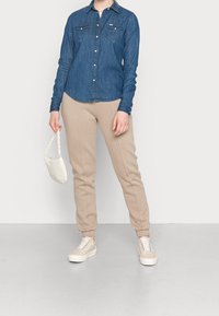 Denim shirt with long sleeves and two chest pockets, paired with beige sweatpants and white sneakers. White bag with a chain strap.