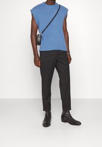 Homme portant un pull sans manches bleu en maille, un pantalon noir sur mesure, des bottines en cuir noir et un sac bandoulière sur fond blanc uni.