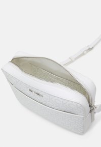 Hvid crossover taske med en struktureret, mønstret overflade. Har en sølvfarvet lynlås, justerbar rem og et rummeligt interiør med logo-foring.