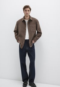 Braunes Cordhemd mit Brusttasche und Knopfleiste, getragen über einem weißen T-Shirt, kombiniert mit dunkelblauen Jeanshosen und schwarzen Schuhen.