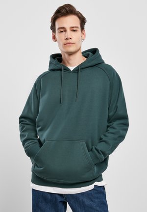 Grön hoodie med framficka, ribbade ärmslut och justerbar dragsko på huvan. Slätt tyg med en avslappnad passform över en vit lagrad skjorta.