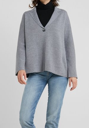 Person trägt eine graue Strickjacke mit einem Knopf über einem schwarzen Rollkragenpullover und hellblauen Jeans, steht vor einem schlichten Hintergrund.