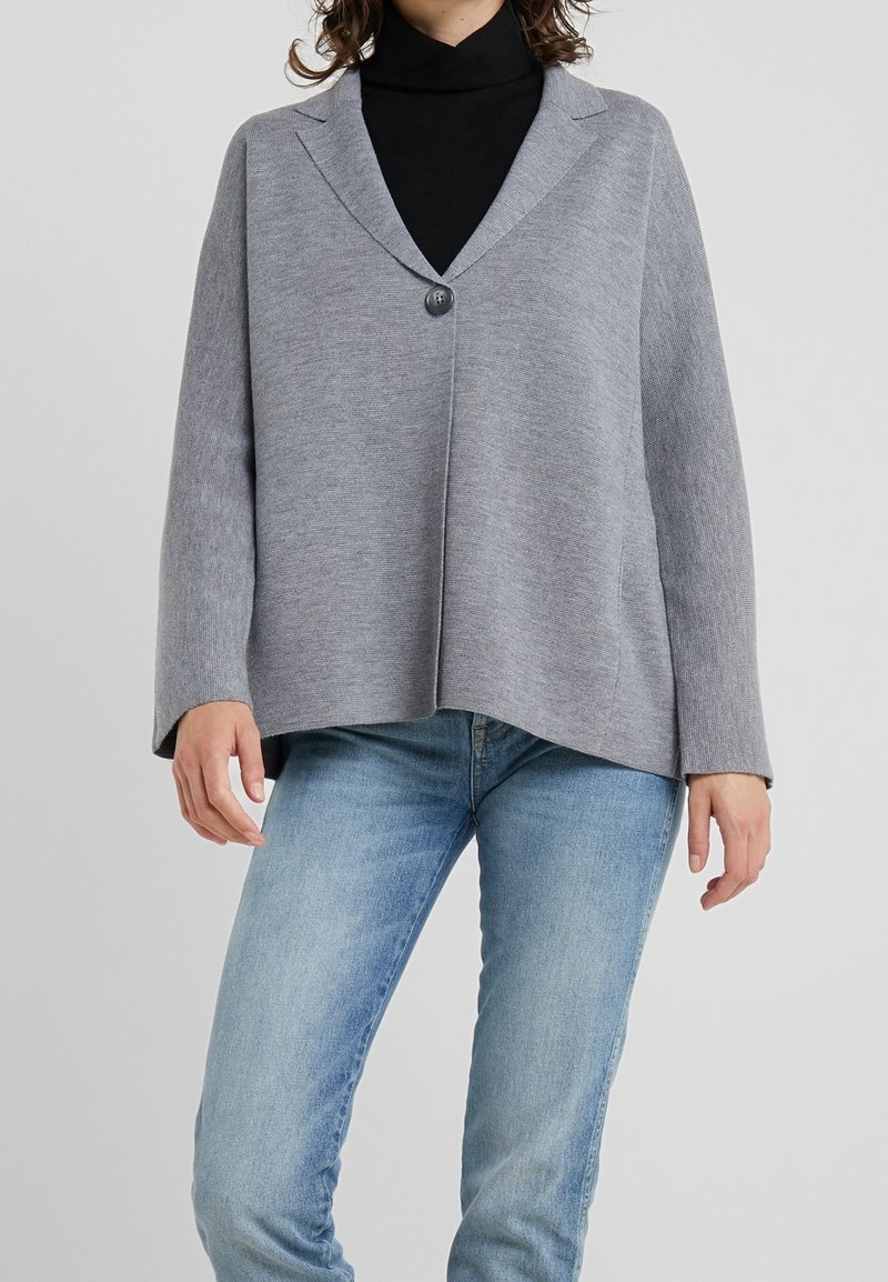 Personne portant un cardigan gris à un bouton par-dessus un col roulé noir avec un jean bleu clair, debout devant un fond uni.