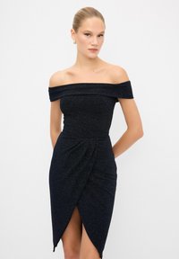 Abito aderente scollato sulle spalle in un blu scuro con tessuto testurizzato. Presenta un design avvolgente e uno spacco laterale per un tocco di stile in più.