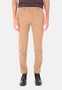 Uomo che indossa pantaloni in velluto a coste beige, scarpe in pelle nera con lacci e una camicia marrone scuro, in piedi contro uno sfondo chiaro.