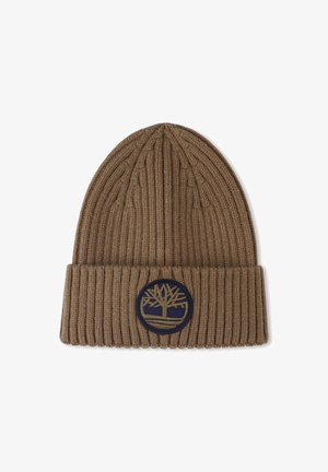 Bruine geribde gebreide beanie met een omgeslagen rand. Heeft een ronde logo patch in marine blauw en goud met een boomontwerp. Zachte textuur.