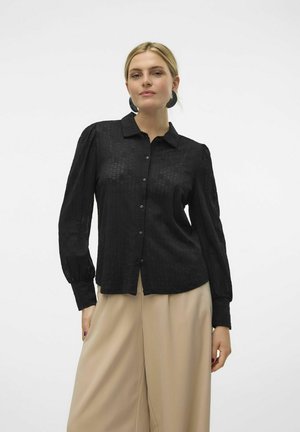 Chemise noire à manches longues à motif texturé, avec un col pointu et des manches bouffantes. Associée à un pantalon large beige.
