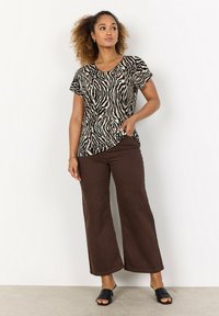 Tiermuster Kurzarmoberteil in Beige und Schwarz, kombiniert mit braunen weit geschnittenen Hosen. Das Model trägt schwarze Riemchensandalen; lässiges Design und Passform.
