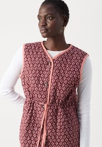 Kvadrātu piegriezuma veste bordo krāsā ar ģeometrisku rakstu, rozā apdari un pogu aizdari. Iekļauta aukliņa jostas vietā, lai pielāgotu fit.