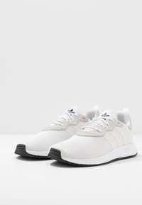 Valkoiset verkkomateriaalista valmistetut tennarit, joissa harmaat mokkanahkaiset yksityiskohdat, kolme valkoista raita, pyöreä kärki ja musta kumipohja.