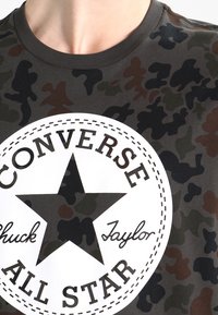 Koszulka w kamuflażowy wzór w ciemnozielonym, brązowym i czarnym kolorze, z dużym białym okrągłym logo z gwiazdą oraz napisem "CONVERSE Chuck Taylor."