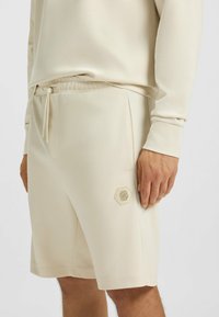 Crème-kleurige shorts van glad, lichtgewicht stof. Voorzien van een trekkoord in de taille en een hexagonale logopatch op de dij.