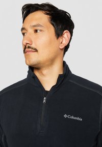 Pull en polaire noir avec un col haut, fermeture éclair et détails de couture subtils. Présente un logo blanc "Columbia" sur la poitrine gauche.