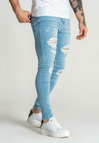Jeans ajustados rasgados de color azul claro, hechos de denim, con parches desgastados y un diseño ceñido, combinados con zapatillas blancas y una camiseta blanca sencilla.